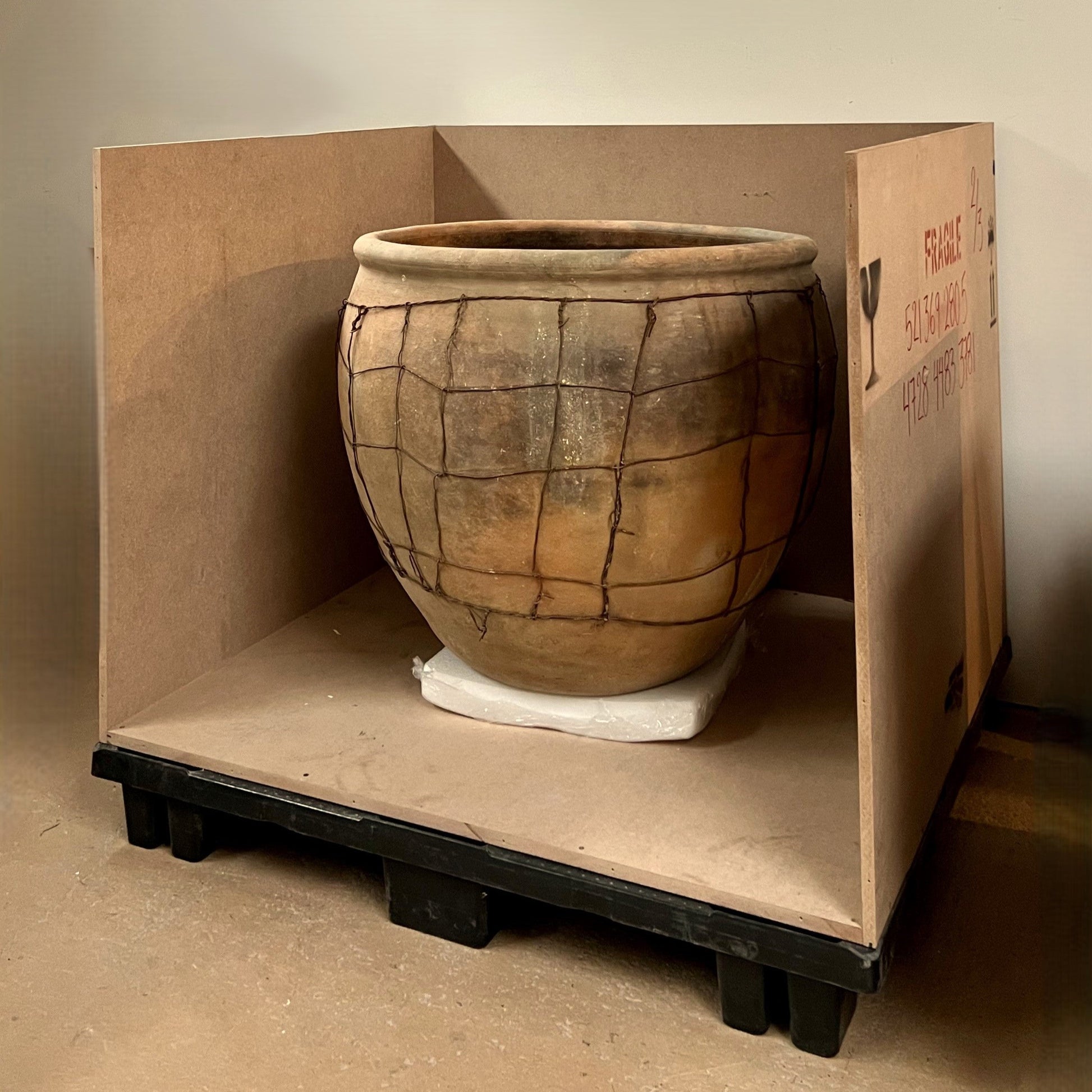 Large ceramic pot inside a crated on a wooden pallet against a plain wall.